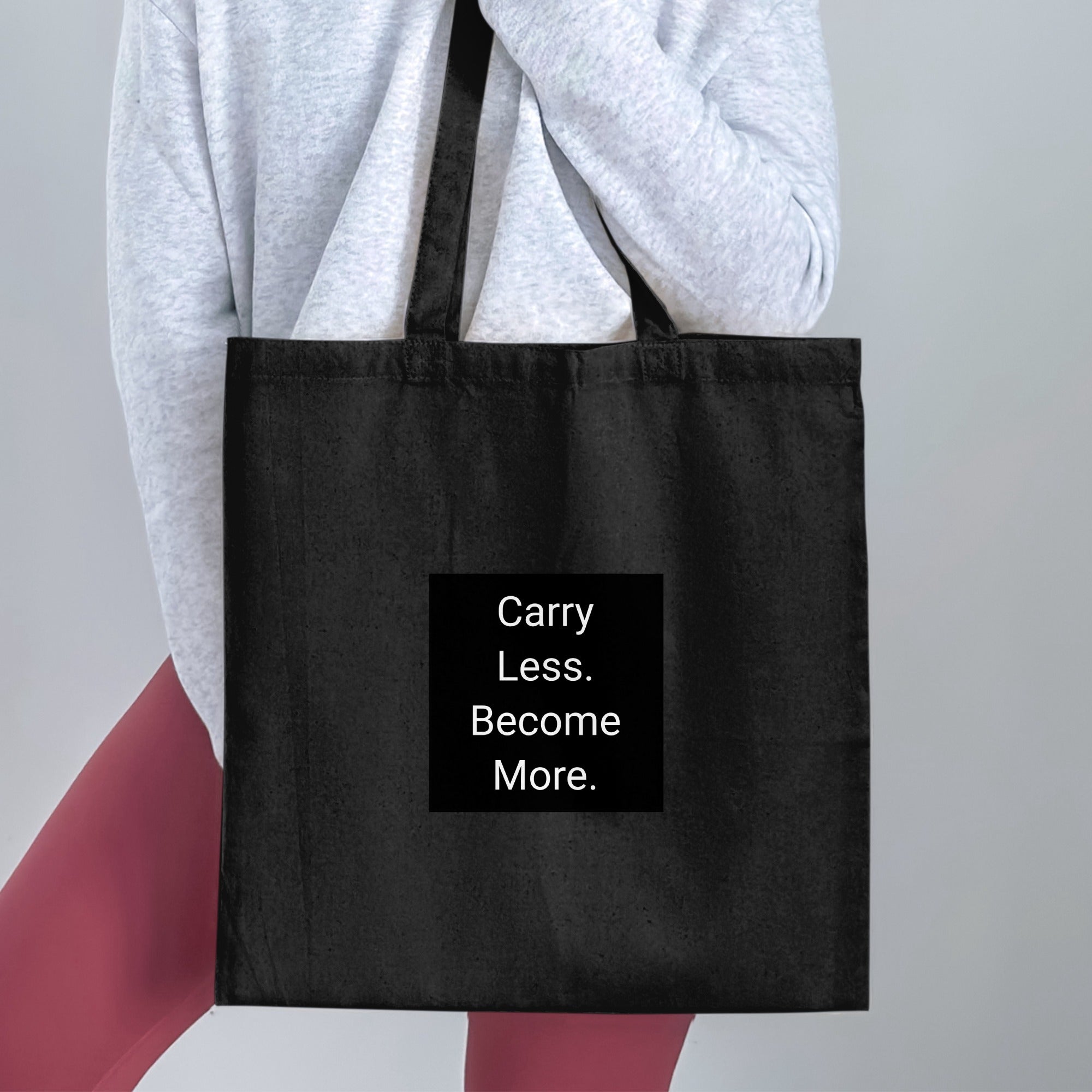 Black tote bag with white text that reads "Carry Less. Become More."