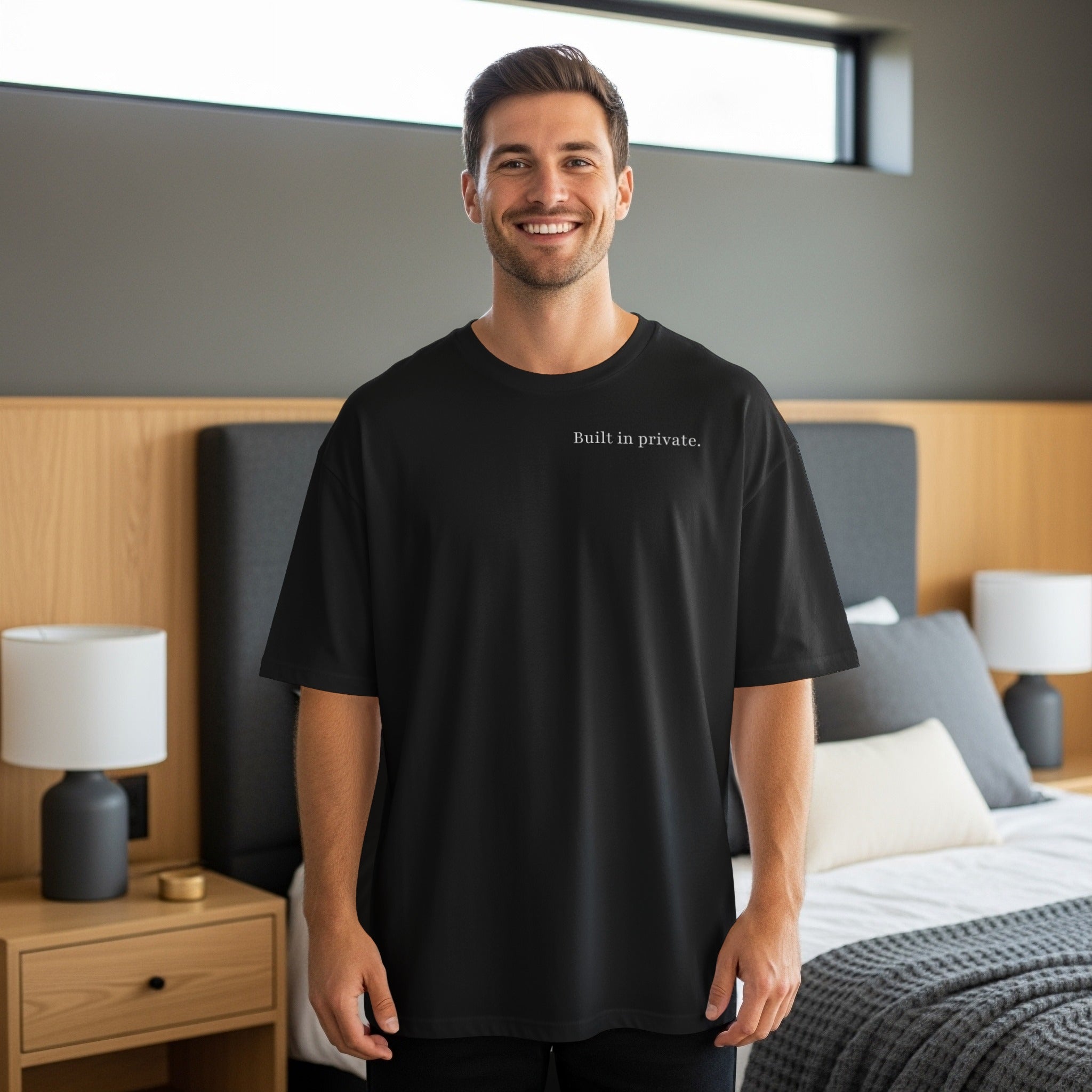 Man in black t-shirt with "Built in private." text, standing in bedroom.