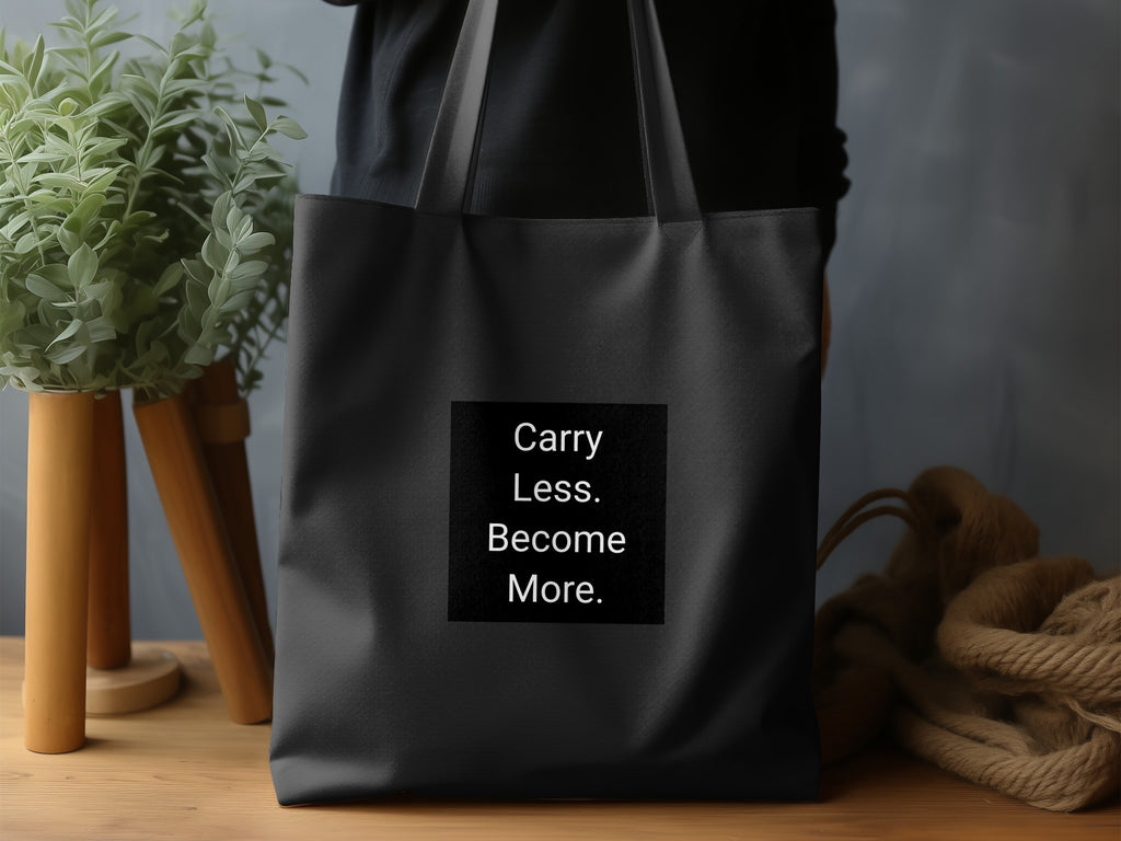Black tote bag with "Carry Less. Become More." text on a black square.